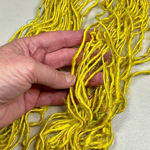 May include: Close-up of bright yellow yarn held in a hand. The yarn is made of many thin strands, with a slightly textured appearance. The color is a vibrant, sunny yellow, and the yarn appears to be soft and flexible.