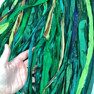 May include: A collection of green fabric strips in various shades, textures, and patterns. The strips are arranged closely together, creating a layered effect. Some strips have a sheen, while others are matte. The color palette ranges from light to dark greens, with hints of blue and yellow.