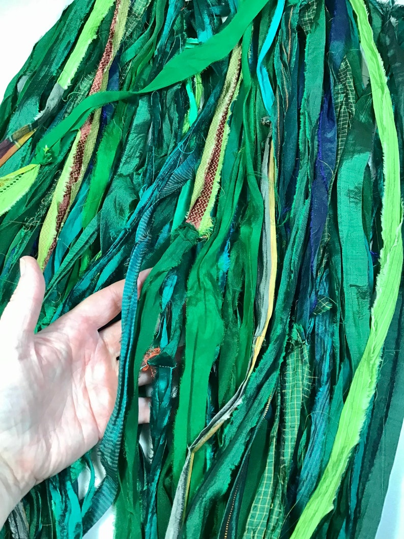 May include: A collection of green fabric strips in various shades, textures, and patterns. The strips are arranged closely together, creating a layered effect. Some strips have a sheen, while others are matte. The color palette ranges from light to dark greens, with hints of blue and yellow.