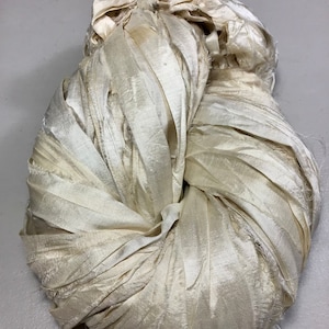 May include: A bundle of off-white, ribbon-like fabric, tightly wound and knotted. The material has a slightly iridescent sheen, with visible frayed edges. The overall shape is rounded, suggesting a decorative or craft element.