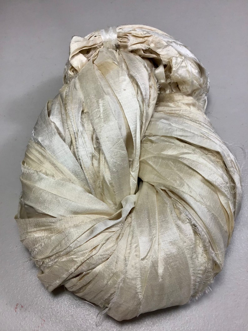 May include: A bundle of off-white, ribbon-like fabric, tightly wound and knotted. The material has a slightly iridescent sheen, with visible frayed edges. The overall shape is rounded, suggesting a decorative or craft element.