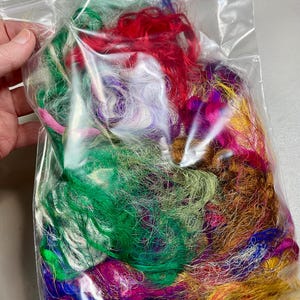 May include: A clear plastic bag filled with colorful craft fibers. The fibers are a mix of green, red, purple, pink, and gold. The bag is held by a hand.
