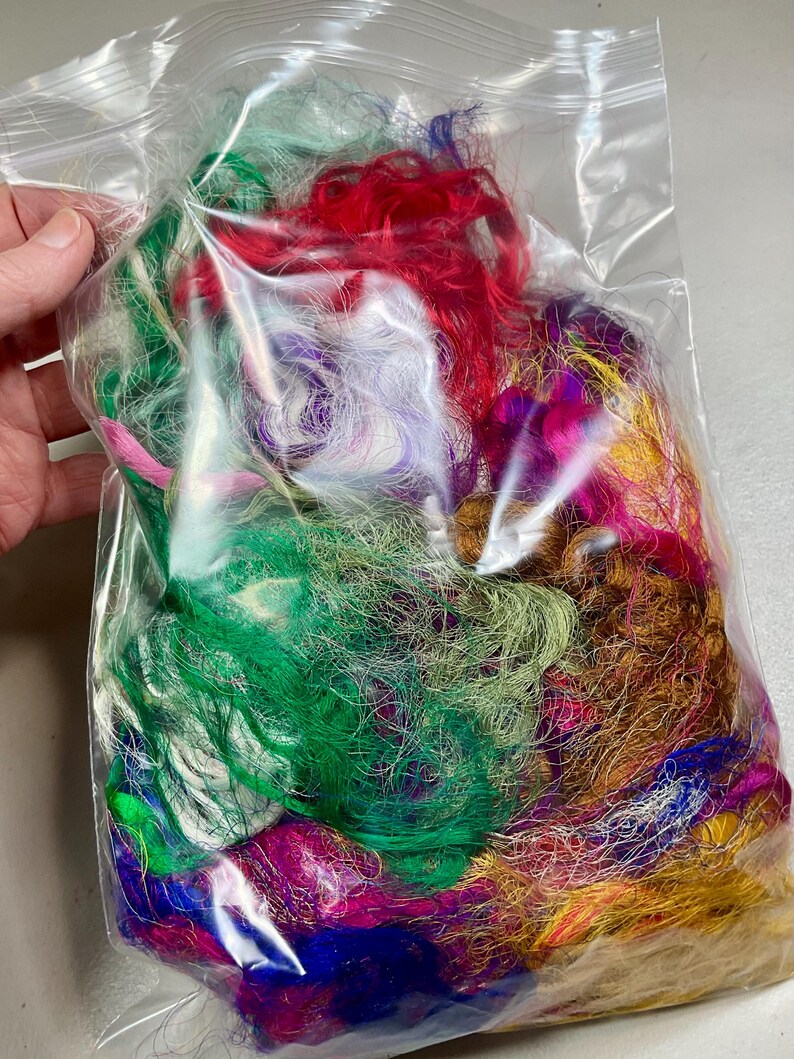 May include: A clear plastic bag filled with colorful craft fibers. The fibers are a mix of green, red, purple, pink, and gold. The bag is held by a hand.