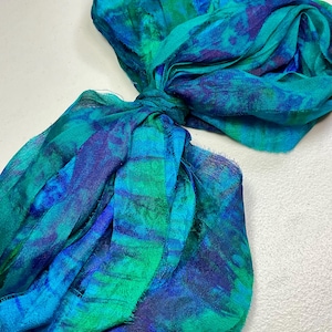 May include: A knotted scarf in shades of teal, blue, and green. The fabric has a watercolor-like pattern with darker blue and purple accents. The scarf is made of a lightweight, flowing material.