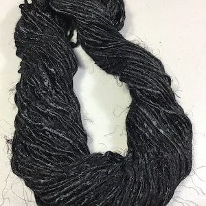 May include: A skein of black yarn with a textured, slightly irregular appearance. The yarn features a deep, dark color with subtle variations in thickness and loose fibers throughout, creating a rustic and hand-spun look for crafting projects.