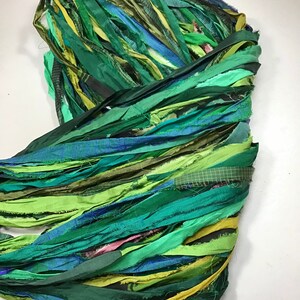May include: A bundle of colorful fabric strips in shades of green, blue, and yellow. The strips are layered and appear to be made of silk or a similar material. The arrangement suggests a craft or textile project.