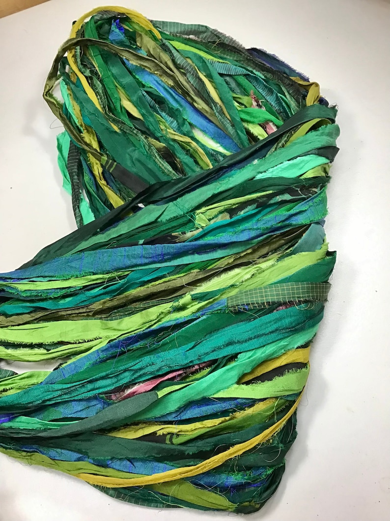 May include: A bundle of colorful fabric strips in shades of green, blue, and yellow. The strips are layered and appear to be made of silk or a similar material. The arrangement suggests a craft or textile project.