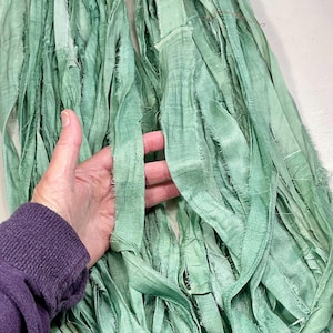 May include: Seafoam green silk ribbon bundles with frayed edges. The ribbons are held in a hand, showing the texture and color variations. Ideal for crafting, scrapbooking, or adding a touch of elegance to gifts.