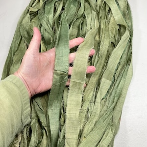May include: Close-up of a bundle of sage green silk ribbon. The ribbon is made of long, thin strips of fabric with a frayed edge. The ribbon is being held by a person wearing a green sleeve.