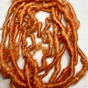 May include: A collection of orange-brown synthetic dreadlocks, arranged in a circular pattern. The dreadlocks vary in thickness and length, with a textured, curly appearance. The color is a warm, earthy tone, suggesting a natural or dyed aesthetic.