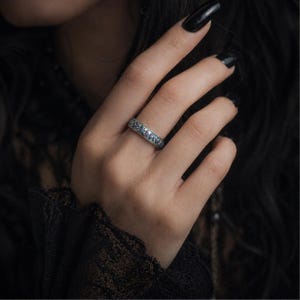 Peut inclure: Une bague en argent ornée de petites pierres bleues rondes. La bague est portée au doigt, avec du vernis à ongles noir et des vêtements sombres en arrière-plan. La bague semble être une alliance ou une bague de mode.