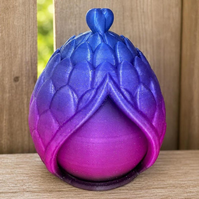 May include: A 3D printed egg-shaped object featuring a gradient color transition from blue at the top to purple and pink at the bottom. The exterior is covered in a textured, overlapping scale pattern with a small heart-shaped finial at the apex.