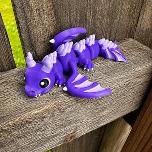 May include: A 3D printed articulated dragon figurine in a vibrant purple color with lighter lavender accents on the horns, wing tips, and back ridges. The creature features large black and white eyes, a segmented body, and detailed wing structures.