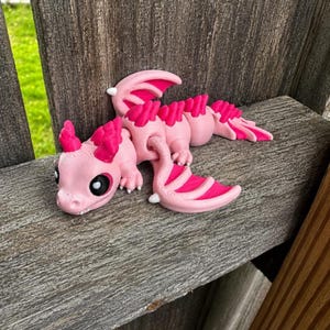 May include: A 3D printed articulated dragon figurine featuring a light pink body with vibrant hot pink accents on the wings, head crest, and back spines. The creature has large black eyes with white highlights and a segmented, flexible body design.