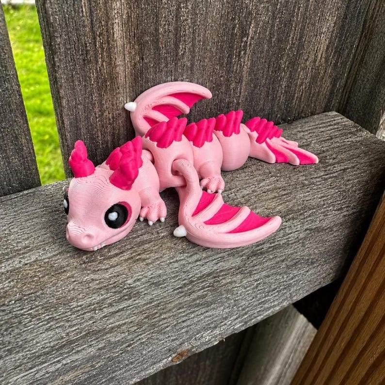 May include: A 3D printed articulated dragon figurine featuring a light pink body with vibrant hot pink accents on the wings, head crest, and back spines. The creature has large black eyes with white highlights and a segmented, flexible body design.