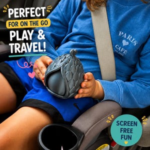 May include: A dark gray, egg-shaped object with a textured, scale-like exterior pattern. The object is held in place, featuring a small heart-shaped detail at the top. Text on the image reads: PERFECT FOR ON THE GO PLAY & TRAVEL! and SCREEN FREE FUN.