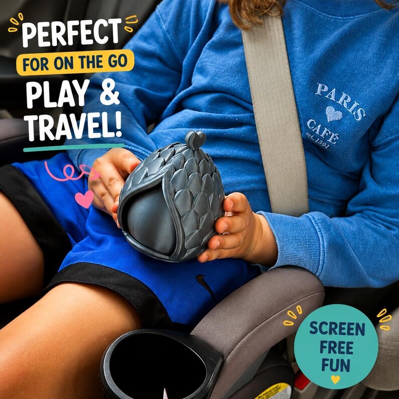 May include: A dark gray, egg-shaped object with a textured, scale-like exterior pattern. The object is held in place, featuring a small heart-shaped detail at the top. Text on the image reads: PERFECT FOR ON THE GO PLAY & TRAVEL! and SCREEN FREE FUN.