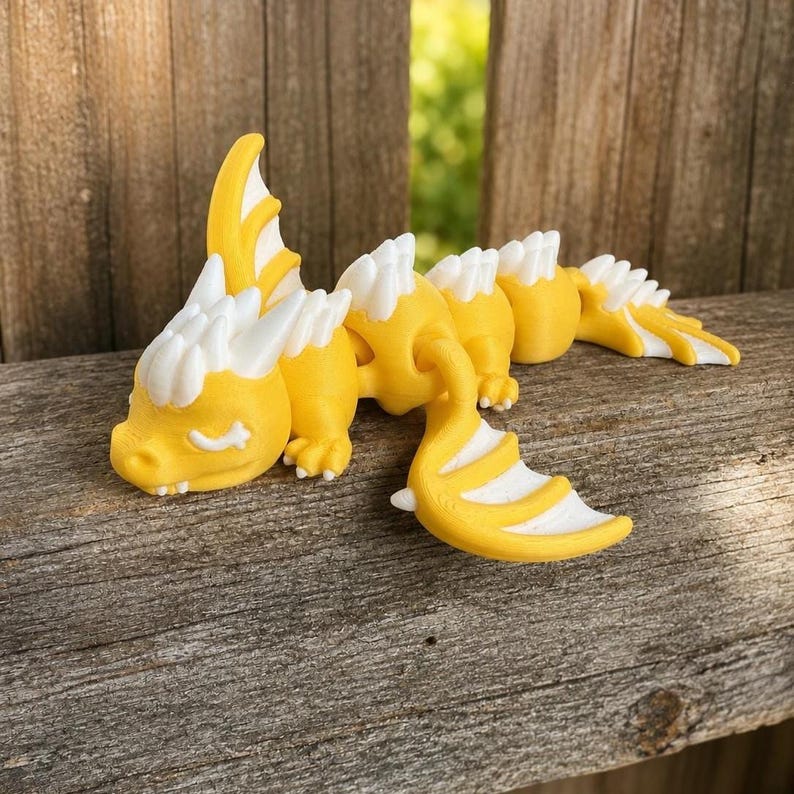 May include: A yellow and white articulated dragon figurine featuring a segmented body, a head with closed eyes, and wings. The creature has a smooth, matte finish with white spikes running along its back and tail, designed with multiple joints.