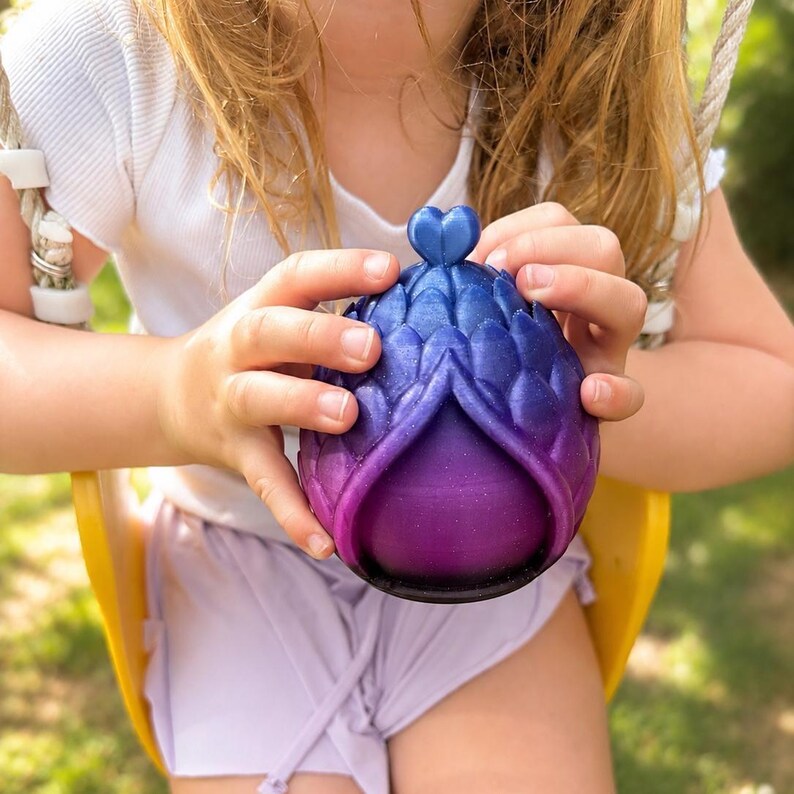 May include: A spherical object with a textured, scale-like exterior that transitions in color from deep blue at the top to vibrant purple and black at the base. The object features a small heart-shaped finial at the apex and a hollow opening.