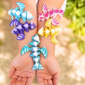 May include: Five articulated dragon figurines in light blue, purple, pink, yellow, and teal colors. Each dragon features a segmented body with white accents on the wings, head, and spine, designed with flexible joints for movement and posing.