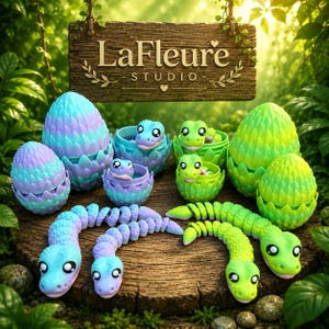 May include: A collection of articulated dragon figures and eggs in blue-purple and green gradients. The figures feature large eyes and textured scales. A wooden sign above reads LaFleur STUDIO with decorative leaf accents and small hearts.