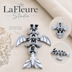 May include: A gray and white articulated dragon toy featuring black horns and the text NAME printed on its segmented body. The dragon has wings and a tail, with additional circular inset views showing the toy from different angles.