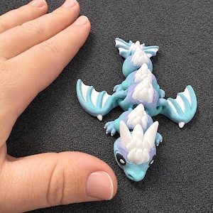 May include: A small articulated dragon figurine featuring a gradient of light blue and purple colors with white spikes along its back and head. The dragon has two wings, a segmented body, and large black eyes, showcasing a 3D printed texture.