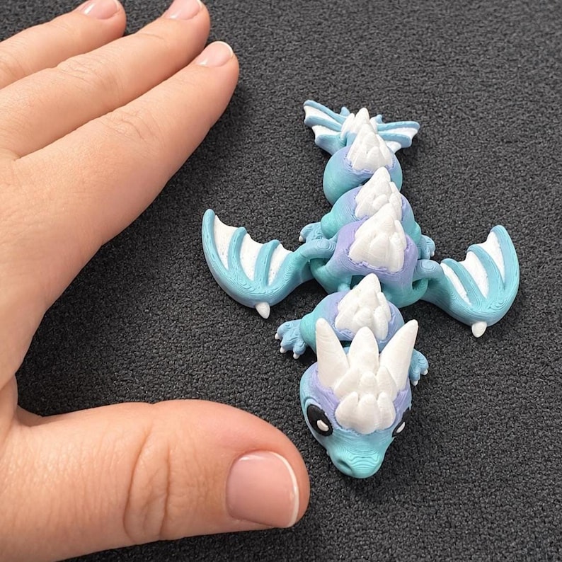 May include: A small articulated dragon figurine featuring a gradient of light blue and purple colors with white spikes along its back and head. The dragon has two wings, a segmented body, and large black eyes, showcasing a 3D printed texture.