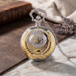 Puede incluir: Reloj de bolsillo plateado y dorado con cadena. La esfera del reloj presenta un globo terráqueo con un emblema de martillo y hoz, rodeado de espigas de trigo y un diseño de sol naciente. El reloj tiene una parte superior decorativa.