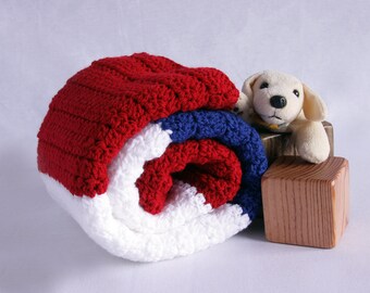 May include: A red, white, and blue crocheted baby blanket rolled up with a stuffed dog toy on top of a wooden block.