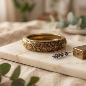 Könnte beinhalten: Ein goldfarbenes Armreif-Armband mit aufwendigen Blumenmustern. Das Armband liegt auf einer weißen Marmoroberfläche. Eine kleine, goldfarbene Schachtel und ein Lavendelzweig sind ebenfalls vorhanden.