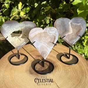 May include: Three heart-shaped agate slices with crystalline centres, each mounted on a black metal wire stand. The stones feature translucent grey and white banding with textured, sparkling druzy cavities. The text THE CELESTIAL REVERIE is visible.