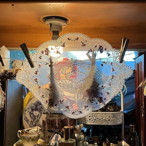 May include: An oval white lace doily featuring intricate floral embroidery with small red and green accents. The lace is suspended by two wooden clothespins on a thin string, showcasing a delicate openwork pattern and scalloped edges.