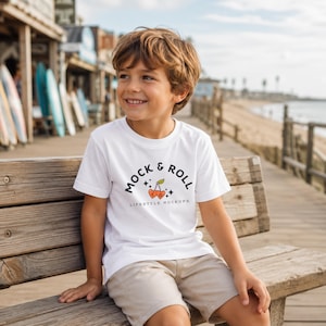 May include: A young person wearing a white t-shirt with the words "MOCK & ROLL" and a cherry graphic. The person is sitting on a wooden bench, with a beach and surfboards in the background. The shirt is a crew neck style.