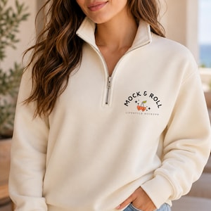May include: A cream-colored quarter-zip sweatshirt featuring a silver metal zipper and a graphic on the left chest. The graphic displays the text MOCK & ROLL LIFESTYLE MOCKUPS in black, accompanied by an illustration of two red heart-shaped cherries.