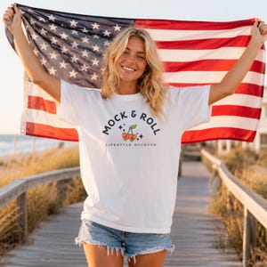 Op de afbeelding: Wit T-shirt met de tekst "MOCK & ROLL" en een kersengrafiek. De persoon houdt een Amerikaanse vlag vast. Het shirt wordt gecombineerd met een spijkerbroek. De achtergrond omvat een houten loopbrug en de oceaan.