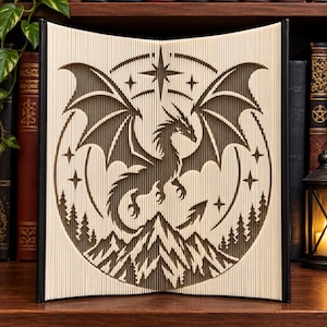 May include: A folded book art sculpture featuring a detailed cutout of a dragon with spread wings, positioned above a mountain range and pine forest. The design is framed by a circular border with stars, created through precise page folding techniques.