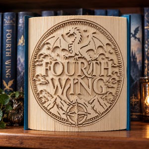 Könnte beinhalten: Eine Buchfaltkunst-Skulptur mit dem Text Fourth Wing. Die Seiten sind kunstvoll gefaltet, um ein kreisförmiges Design zu erzeugen, das eine Drachensilhouette, Wolken, Berggipfel und eine Windrose enthält, die mittig auf den Buchseiten liegen.