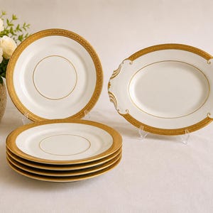 Może przedstawiać: Zestaw białych porcelanowych naczyń do serwowania ze złotymi obramowaniami i akcentami. Zestaw zawiera okrągłe talerze i owalny półmisek. Talerze są ułożone w stos, a półmisek ma dekoracyjne uchwyty.