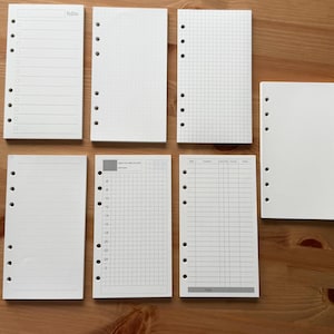 May include: Assortment of white planner inserts with various layouts, including to-do lists, dot grids, graph paper, lined pages, and daily schedules. Each insert has pre-punched holes for binding. The inserts are arranged on a wooden surface.