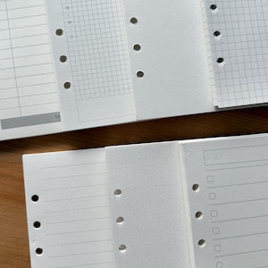 May include: A close-up shot of several white planner inserts with pre-punched holes for a binder. The inserts feature various layouts, including lined pages, grid paper, and a time-based schedule. The pages are clean and ready for use.