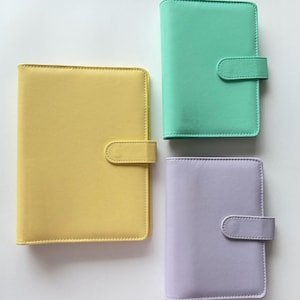 May include: Four colorful, leather-like notebooks in yellow, mint green, and lavender. Each notebook has a rounded flap closure. The notebooks are arranged on a white surface.