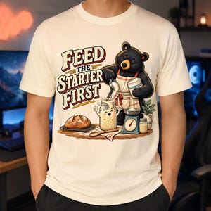May include: A cream-colored t-shirt featuring a graphic of a black bear wearing an apron, pouring flour into a smiling glass jar. The text reads "FEED THE STARTER FIRST" in bold letters. A loaf of bread, a kitchen scale, and a small jar are also shown.