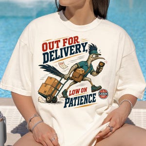Può includere: Una t-shirt color crema con la grafica di un roadrunner in uniforme blu, che trasporta un pacco marrone e tira un carrello a mano con due scatole. Il testo recita "OUT FOR DELIVERY, LOW ON PATIENCE" e "LABOR DAY".