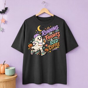 Puede incluir: Una camiseta negra con el gráfico de un fantasma de dibujos animados que lleva una gorra de béisbol morada y zapatillas deportivas, sosteniendo un café helado. El texto dice "RUNNING ON FRIGHTS & ICED COFFEE" en letras coloridas, rodeado de calabazas, una luna creciente y hojas.