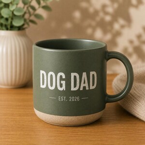 May include: A green ceramic mug with the words "DOG DAD" in white block letters. Below the text is "EST. 2026". The bottom of the mug is a natural beige color. The mug has a curved handle.