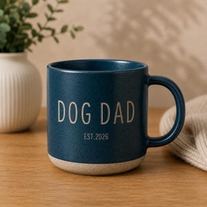 May include: A speckled, navy blue ceramic mug with a beige base. The mug features the text "DOG DAD" in white, with "EST. 2026" below. The mug has a large, curved handle.