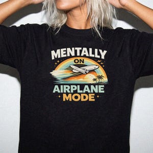 Puede incluir: Camiseta negra con el texto "MENTALLY ON AIRPLANE MODE" sobre un gr&aacute;fico de un avi&oacute;n, arco&iacute;ris, oc&eacute;ano y palmeras. El dise&ntilde;o utiliza colores azul, naranja y verde.
