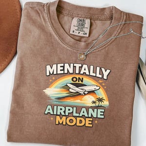 Puede incluir: Una camiseta marr&oacute;n Comfort Colors con el texto "MENTALLY ON AIRPLANE MODE" en blanco, naranja y azul. El gr&aacute;fico incluye un arco&iacute;ris, un avi&oacute;n y palmeras. Un collar plateado con un colgante est&aacute; sobre la camiseta.