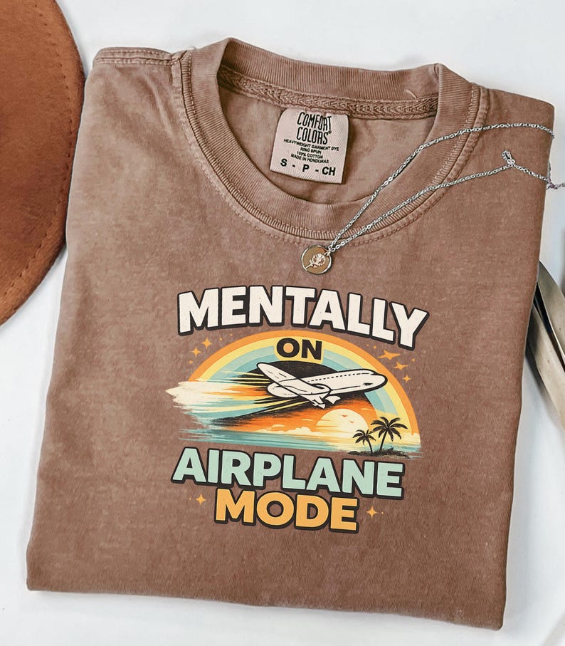 Puede incluir: Una camiseta marr&oacute;n Comfort Colors con el texto "MENTALLY ON AIRPLANE MODE" en blanco, naranja y azul. El gr&aacute;fico incluye un arco&iacute;ris, un avi&oacute;n y palmeras. Un collar plateado con un colgante est&aacute; sobre la camiseta.